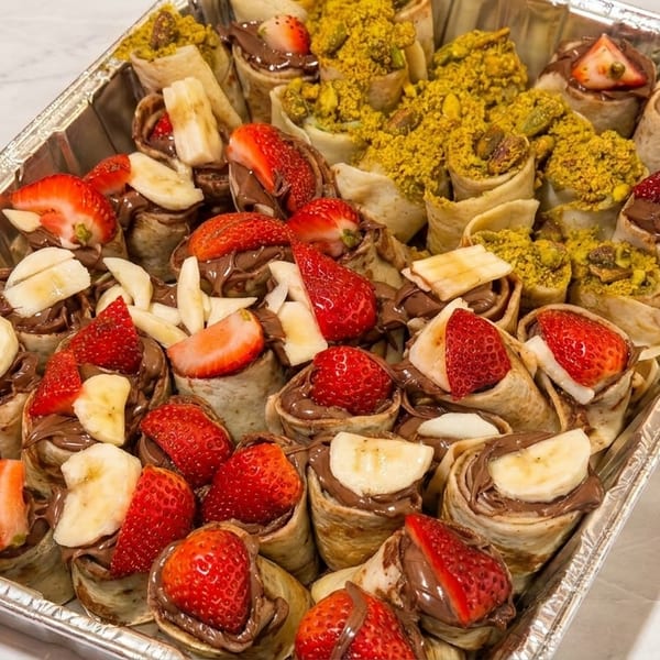 A catering tray filled with dessert wraps, some with strawberries and bananas, others with pistachios.